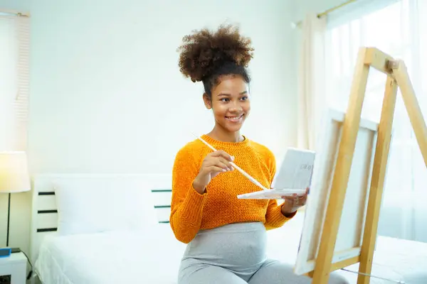 Happy cheerful American - African black ethnicity pregnant woman enjoy drawing and painting a water color on canvas in bedroom, hobby and activity for pregnant woman.