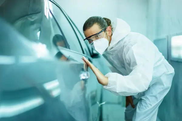 Professional automotive painting technician in chemical protecting suit inspecting car painting quality in painting chamber together. Car painting and detailing technicians portrait.