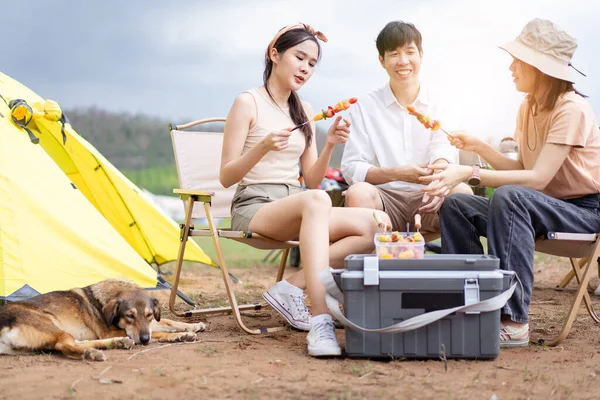 A group of Asian people enjoy camping and cooking together, men and women preparing meat to make a bar-b-q or BBQ at their camping site in the forest.