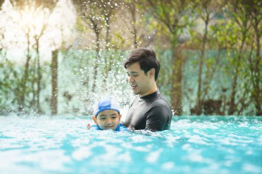 Mutlu Asyalı baba ve oğul yüzme havuzunda birlikte oynayıp yüzüyor. Bir çocuk babasıyla su oynamaktan zevk alıyor. Aile içinde yaz ve tatil aktiviteleri.