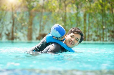 Mutlu Asyalı baba ve oğul yüzme havuzunda birlikte oynayıp yüzüyor. Bir çocuk babasıyla su oynamaktan zevk alıyor. Aile içinde yaz ve tatil aktiviteleri.