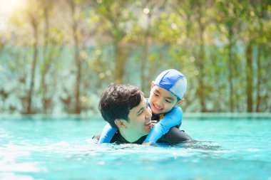 Mutlu Asyalı baba ve oğul yüzme havuzunda birlikte oynayıp yüzüyor. Bir çocuk babasıyla su oynamaktan zevk alıyor. Aile içinde yaz ve tatil aktiviteleri.