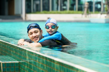 Mutlu Asyalı baba ve oğul yüzme havuzunda birlikte oynayıp yüzüyor. Bir çocuk babasıyla su oynamaktan zevk alıyor. Aile içinde yaz ve tatil aktiviteleri.