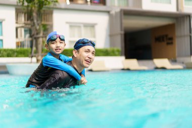 Mutlu Asyalı baba ve oğul yüzme havuzunda birlikte oynayıp yüzüyor. Bir çocuk babasıyla su oynamaktan zevk alıyor. Aile içinde yaz ve tatil aktiviteleri.