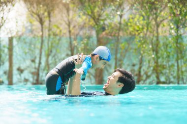 Mutlu Asyalı baba ve oğul yüzme havuzunda birlikte oynayıp yüzüyor. Bir çocuk babasıyla su oynamaktan zevk alıyor. Aile içinde yaz ve tatil aktiviteleri.