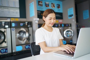 Mutlu Asyalı güzel kadın self servis çamaşır makinesi kullanıyor KIOSK ya da otomatik çamaşır makinesi satıcısı. Çamaşırhanedeki bekleme salonunda dizüstü bilgisayarda çalışan bir kadın..