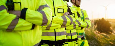 Elektrik mühendislerinin grup portresi arka plandaki günbatımına karşı kameraya poz veriyor. Güvenlik giysisi ve başlık giymiş mühendisler kameraya poz veriyorlar..