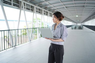 Asyalı genç iş kadınının portresi şehir merkezinde yürürken dizüstü bilgisayar tutuyor ve kullanıyor. Asyalı iş kadını dizüstü bilgisayar kullanıyor.