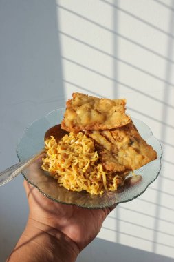 Kahvaltıda menüde kızarmış şehriye ve kızarmış tempeh olan bir yemeğin fotoğrafı beyaz bir duvar arkasında bir adam tarafından tutuluyor.