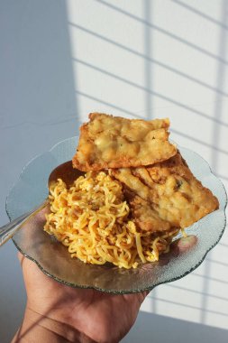 Kahvaltıda menüde kızarmış şehriye ve kızarmış tempeh olan bir yemeğin fotoğrafı beyaz bir duvar arkasında bir adam tarafından tutuluyor.