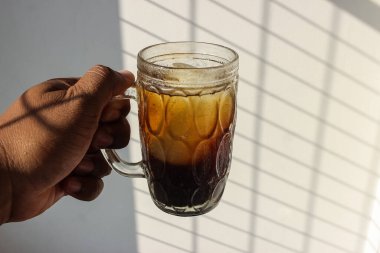 Buzlu bir kahve bardağının fotoğrafı. Büyük bir cam fincanda beyaz bir duvarın arkasında duran bir adam tarafından tutuluyor.