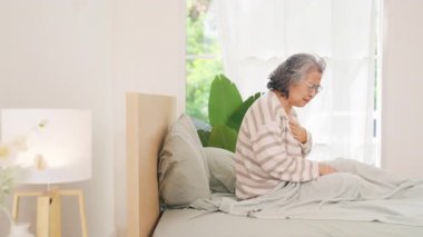 Asyalı olgun bir kadın yatak odasındaki yatakta kalp krizi geçiriyor. Yaşlı kadın göğüs bölgesinde ağrı ve nefes darlığı çekiyor. Acil sağlık ve sağlık sigortası kavramı.