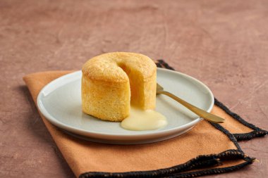 Tek bir vanilyalı lav kekinin, beyaz bir tabağa sızan erimiş, zengin bir merkezi olan yakın çekimi. Pasta, dokulu bir peçetede altın bir çatalla servis edilir. Yumuşak ve havadar bir dış görünüşü ve pürüzsüz, kremsi bir dolgusu vardır..