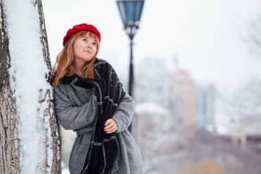 Kızıl saçlı genç kadın, kırmızı şapkalı, karlı parkta yürüyor. 