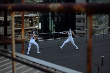 Arka planda şehir varken çatıda çalışan iki eskrimci kadın.