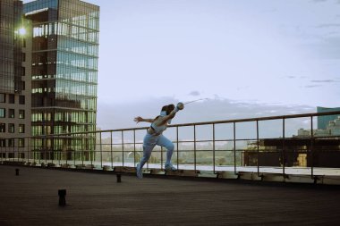 Arka planda şehir varken çatıda eskrim antrenmanı yapan kadın. 