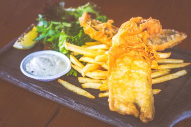 Bira ezilmiş balık ve patates kızartması, salata ve tartar sosuyla ahşap tabakta servis edilir.