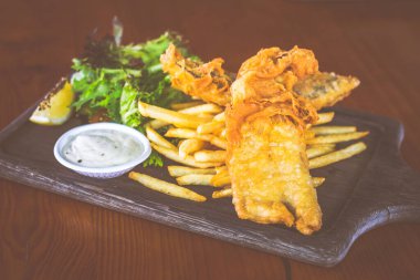 Bira ezilmiş balık ve patates kızartması, salata ve tartar sosuyla ahşap tabakta servis edilir.