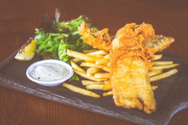 Bira ezilmiş balık ve patates kızartması, salata ve tartar sosuyla ahşap tabakta servis edilir.