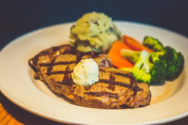 340 gramlık New York bifteği, ızgara ve üzeri bitki yağı ile kaplı. Yanında patates püresi ve taze sebzeyle servis edilir.