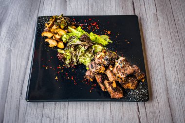 Siyah tabakta servis edilen salata ve sotelenmiş mantarlı biftek küpü.