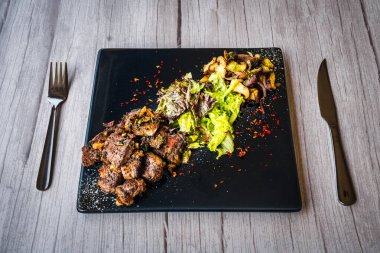 Siyah tabakta servis edilen salata ve sotelenmiş mantarlı biftek küpü.