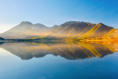 Lake and mountains in a valley at dawn. Reflections on the surface of the lake. Mountain landscape at sunrise. Foggy morning. Natural landscape with bright sunshine.