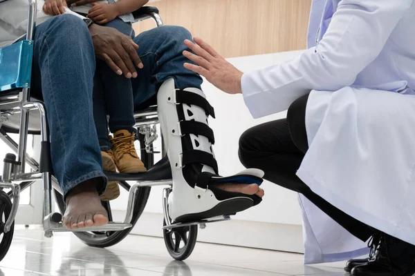 Doctors examines a black patient who has had an accident with a broken leg accompanied by his wife and child.
