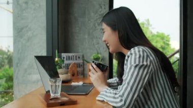 Genç Asyalı kadın kahve dükkanında oturuyor ve arkadaşlarıyla laptopla konuşuyor ve kahve içiyor..