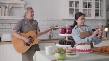 Mutlu son sınıf Asyalı çift evde gitar çalıp kek pişiriyor.