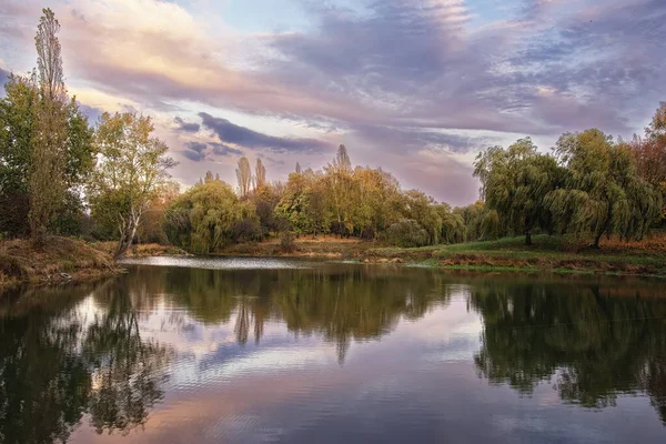 stock image Summer landscape - sunset over the river bank with trees and bushes. The sky with clouds is reflected in the water.