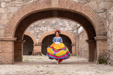 Frida 'dan esinlenerek Frida Kahlo' yu andıran Meksikalı bir kadın narin kemer yolları altında yaşlanmış bir sandalyeyi şereflendirir. Bir ışık ve gölge oyunuyla yıkandı, geleneksel kıyafetleri ve derin derin bakışları