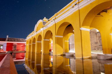 Antigua Guatemala 'daki Tanque La Union' ın gece görüşü bir zamanlar umumi çamaşır alanı olarak kullanılan berrak yıldızlı gökyüzünün altında ikonik sarı koloni kemerleri ve su yansımalarını gösteriyor..
