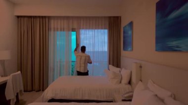 Beyaz çarşaflı ve modern dekorlu rahat ve zarif bir otel odası. Bir adam gün batımında nefes kesen okyanus manzarasını göstermek için perdeleri açar. Kamera pencereye doğru hareket ediyor, deniz ve ufkun panoramik bir sahnesine geçiyor. Mükemmel görüntüler