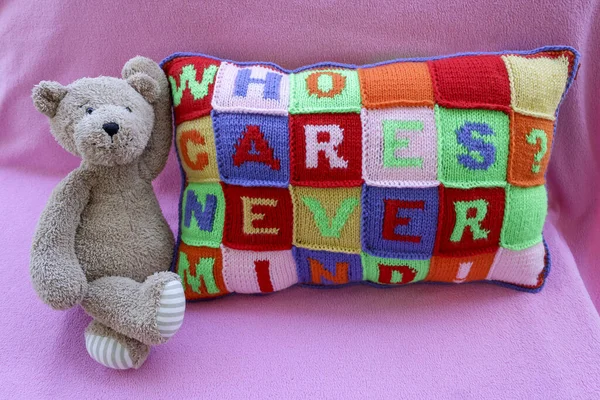 Teddy bear sitting beside hand knitted pillow with multi colored careless phrases