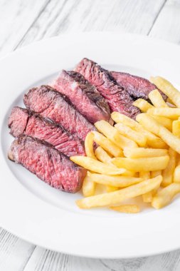 Tahta bir masada beyaz bir tabakta servis edilen orta pişmiş pirzola bifteği ve patates kızartması.