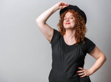 Kırmızı kıvırcık saçlı mutlu kadın gri arka planda poz veriyor, fotokopi uzayı kovboy şapkasını parlatıyor. 