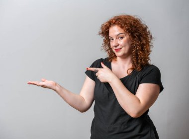 Kırmızı kıvırcık saçlı mutlu kadın gri arka planda poz veriyor, fotokopi alanı elleriyle görünüyor.