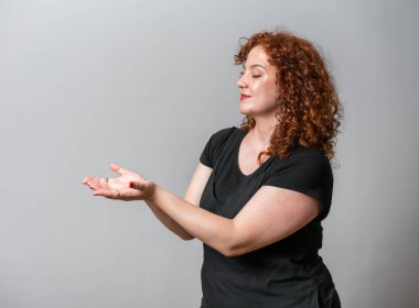 Kırmızı kıvırcık saçlı mutlu kadın gri arka planda poz veriyor, fotokopi alanı elleriyle görünüyor.