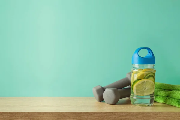 Fitness background with infused water bottle, towel and dumbbells on wooden table over blue background