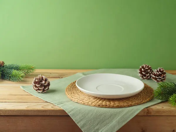 Yeşil duvarın üzerinde masa örtüsü olan boş beyaz tabak ve çam ağacı süsleme arka planı. Tasarım ve ürün gösterimi için Noel modeli.