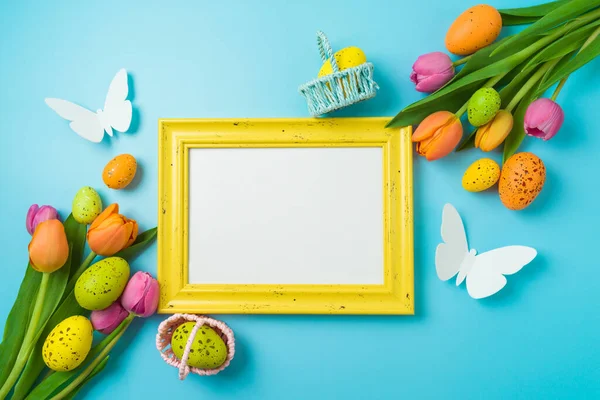 Easter holiday picture frame mock up  with easter eggs and tulip flowers on blue background. Top view, flat lay