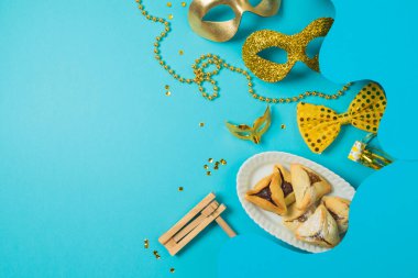 Karnaval maskeli Purim bayramı konsepti, gürültü çıkarıcı ve mavi arka planda hamantaschen kurabiyeleri. Üst görünüm, düz konum