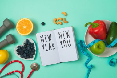 New Year resolutions concept. Healthy lifestyle and balanced diet vegetables, fruits, medical stethoscope, dumbbells and notebook on blue background clipart