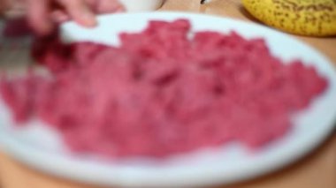 The selective focus highlights the metal spoon as it breaks apart a block of fresh, raw minced beef.