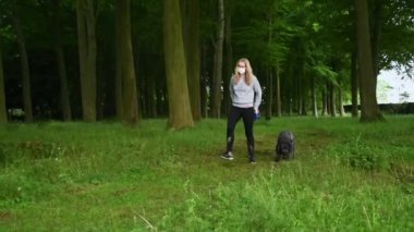Bir kadın yüz maskesi takar ve siyah labrador köpeğini yeşil yapraklı bir orman boyunca kameraya doğru yürütür.