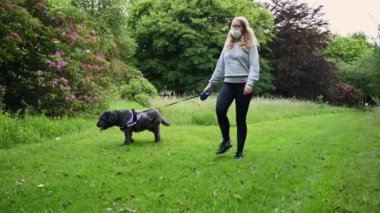 PPE maskeli bir kadın ve siyah bir labrador köpeğini ağaçlarla kaplı bir orman yolunda kameranın önünden geçirir.