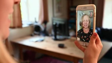 Sarışın bir kadın gülümseyen bir kadınla video görüşmesi yaparken elinde bir akıllı telefon tutarken omzundan dışarı doğru çek.