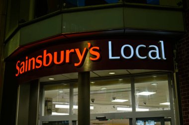 12 Kasım 2022 'de mağaza girişinin üstündeki neon Sainsbury' nin yerel tabelası müşteriler için sıcak ve sıcak bir karşılama parlaklığı sağlıyor..
