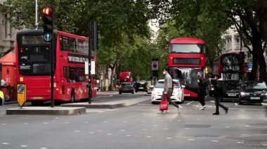 Londra 'da yayalar caddeyi geçerken kırmızı otobüsler geçmekte, PPE maskeli önlemler almakta ve salgın sırasında sorumlu davranışlar sergilemektedirler..